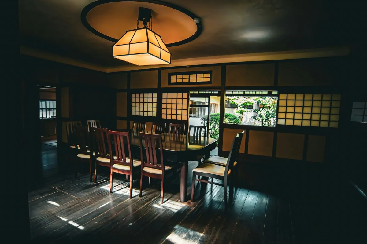 A long dining table under a paper lantern beside garden windows