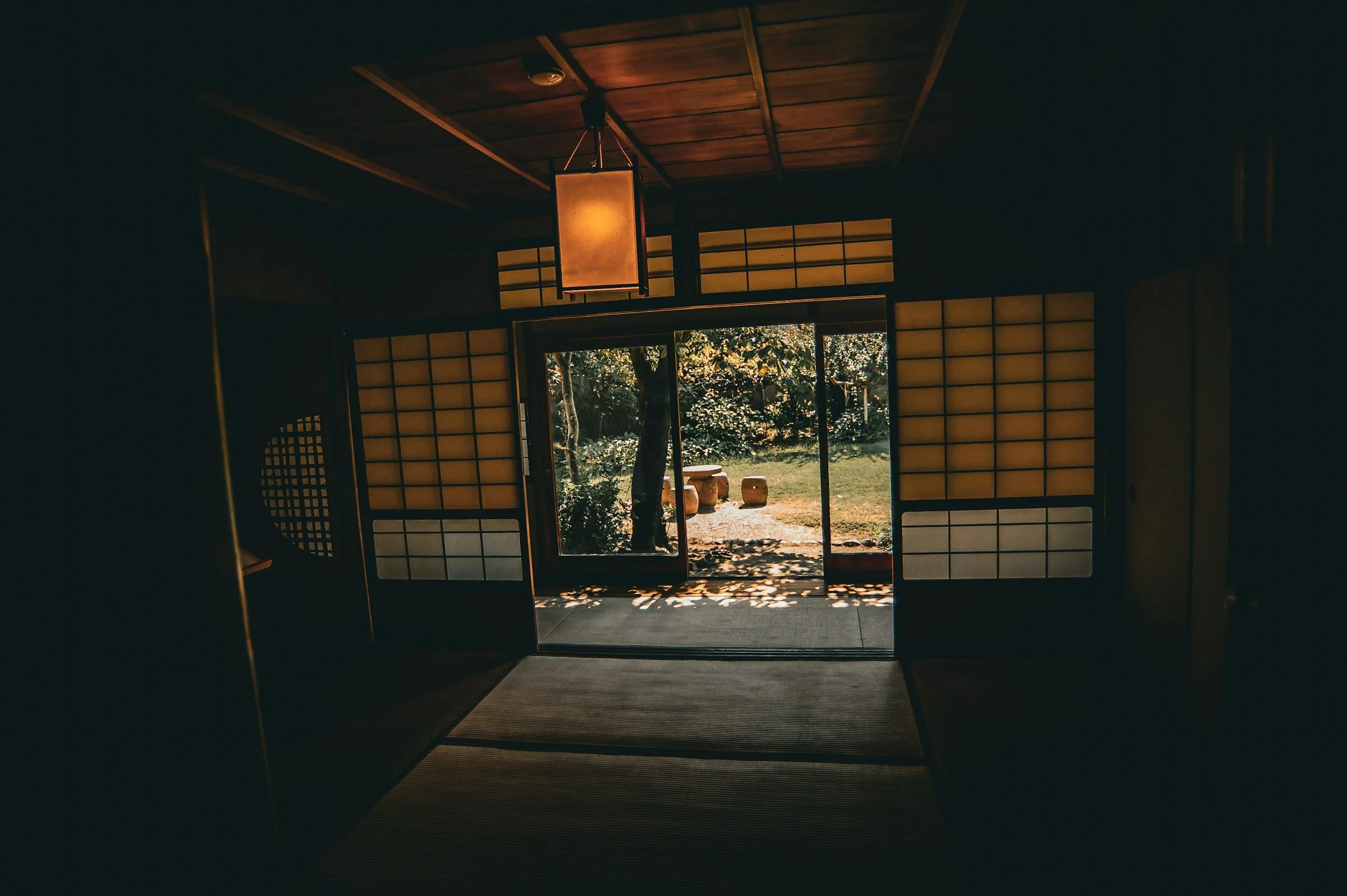 A dark timber room lit by a hanging paper lantern, its doors open onto a sunlit garden