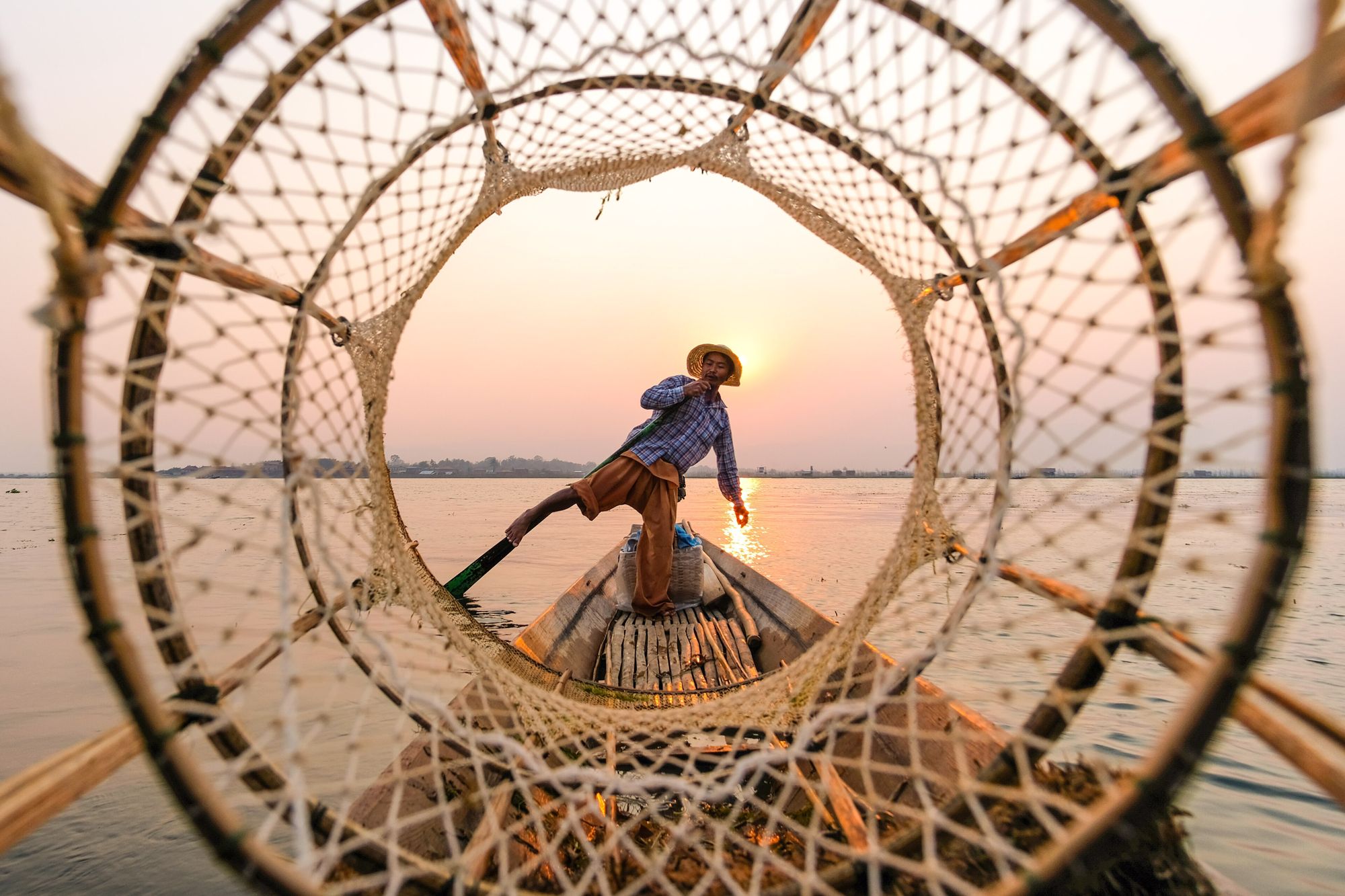 A fisherman on Inle Lake framed through the round mouth of a bamboo fishing net at sunset