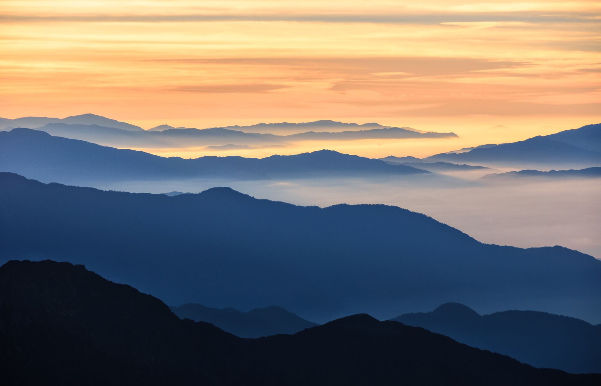 Layers of blue mountain ridges under a pale orange sunrise, mist filling the valleys