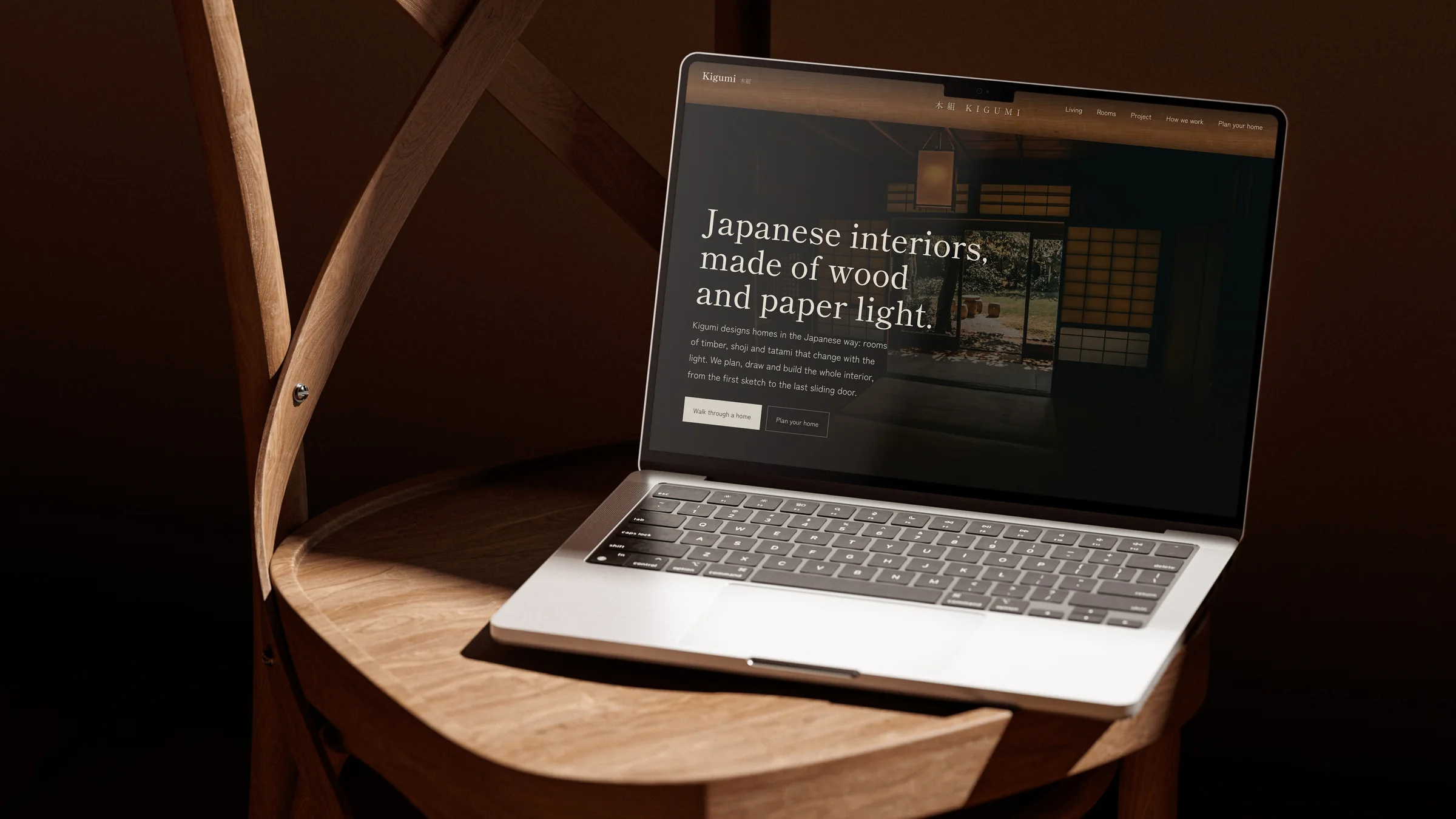 MacBook に表示した Kigumi のトップ。見出し Japanese interiors, made of wood and paper light. 暗い部屋の木の椅子の上